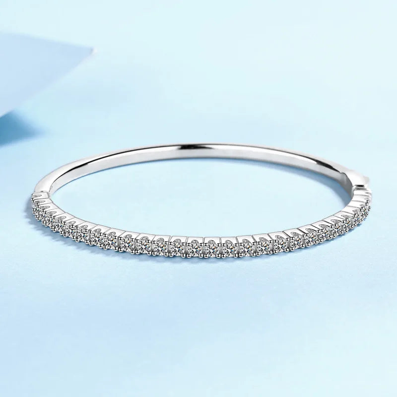 A thin silver-toned bangle bracelet with a row of small clear stones along the front, photographed on a soft light blue background.
