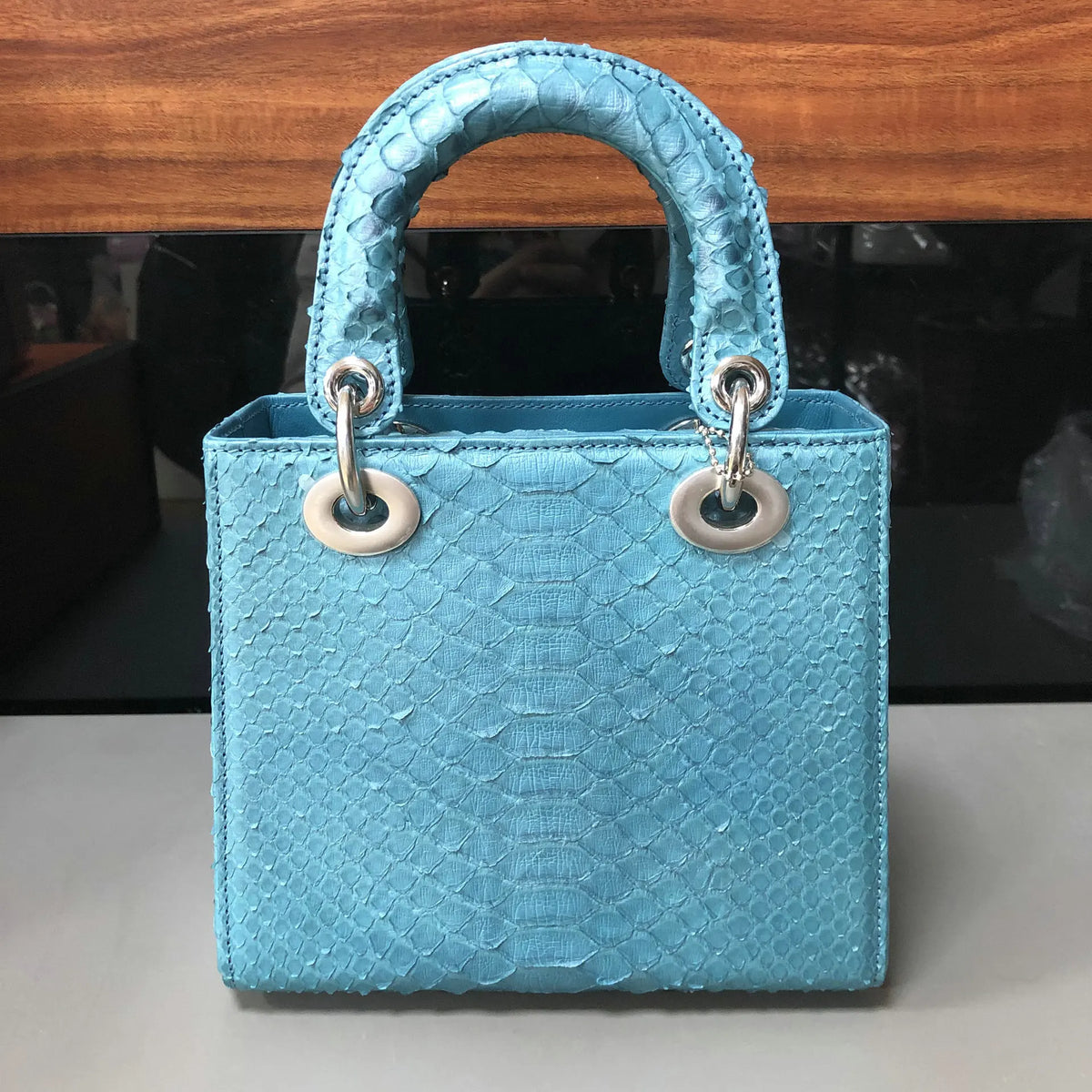 A light blue python-textured leather handbag with a top handle and silver-tone hardware, photographed on a light surface with a dark background.