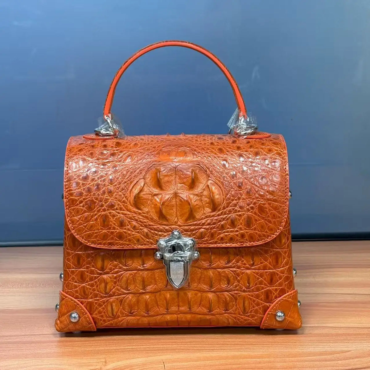 An orange-brown crocodile-embossed leather handbag with a top handle, silver-tone hardware, and a decorative front clasp, displayed on a wooden surface against a blue background.