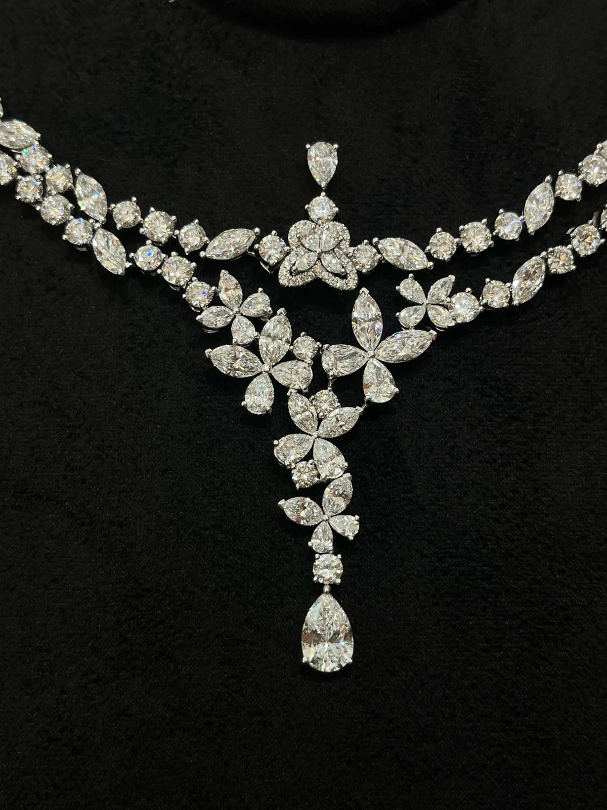 A sparkling silver-tone diamond necklace with a floral and leaf motif, featuring a central teardrop diamond pendant, displayed on a black velvet background.