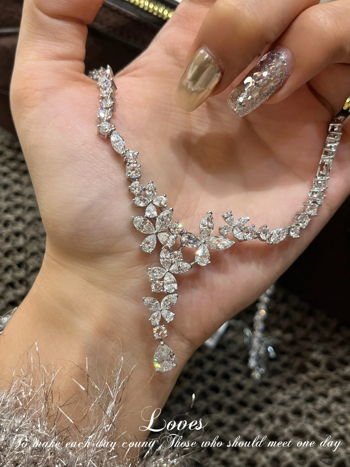 A close-up of a hand holding a sparkling silver-toned floral necklace with clear rhinestones. The necklace features a Y-shaped design with a central cluster of flower motifs and a dangling teardrop crystal at the bottom.
