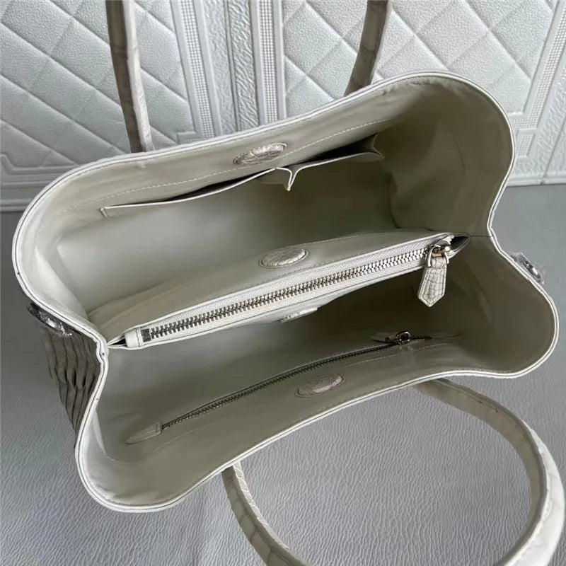 An off-white crocodile-embossed handbag shown from an overhead angle, highlighting its interior lining, zippered compartments, and quilted background.