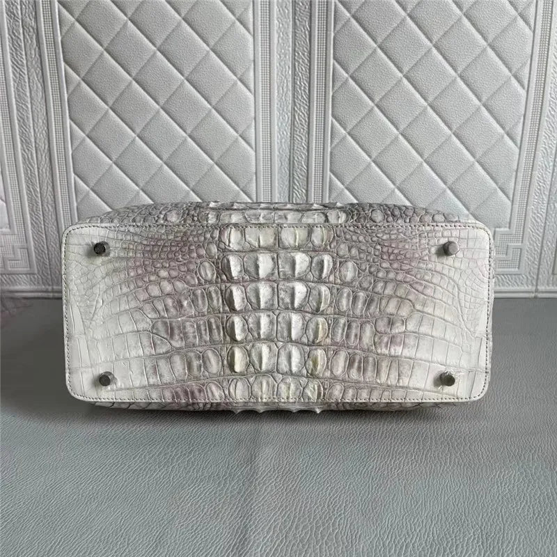 A silver alligator leather handbag shown from the bottom, featuring a textured crocodile pattern and silver metal studs, set against a quilted white leather background.