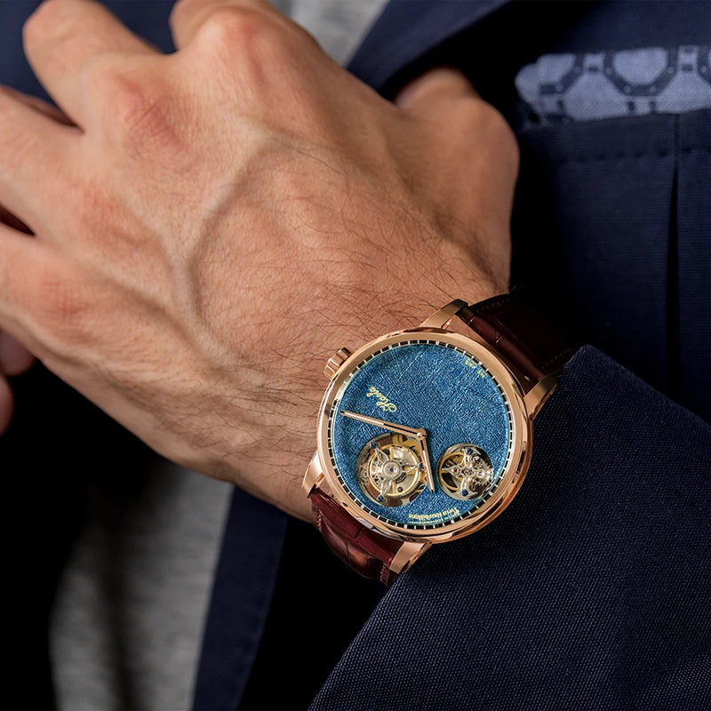 A close-up of a luxury wristwatch with a blue textured dial, rose gold case, and brown leather strap, worn on a man's wrist.