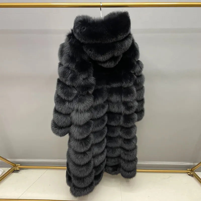 A long black fur coat with a large shawl collar and layered horizontal texture, hanging on a gold clothing rack against a light gray wall.