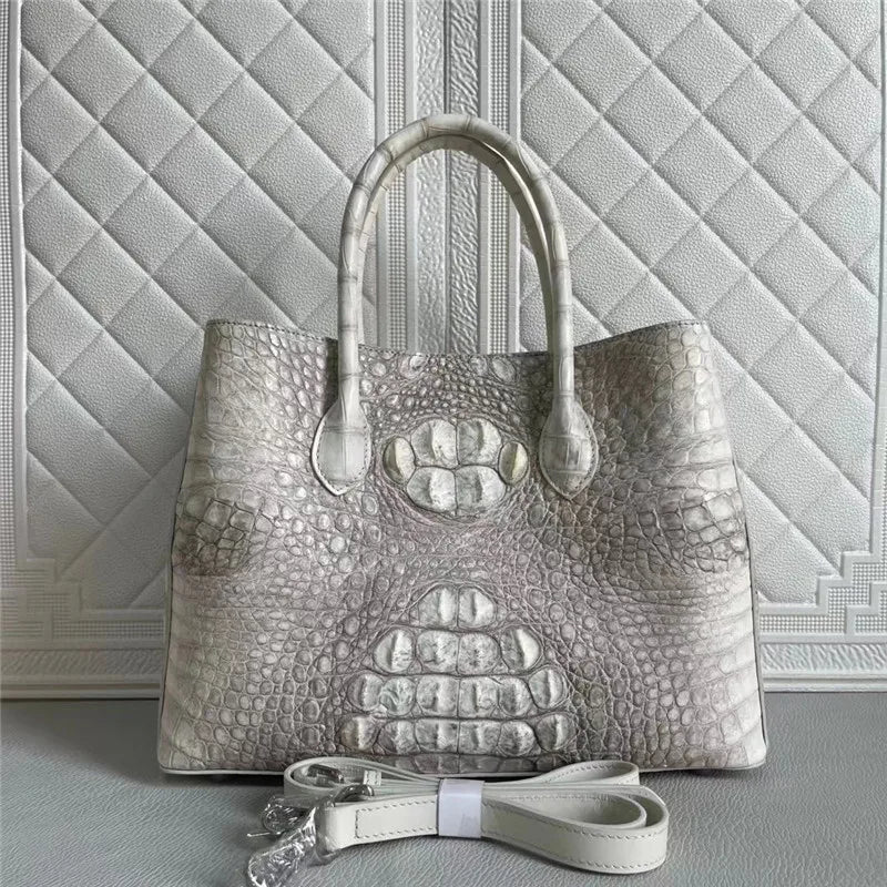 A structured tote handbag with a glossy crocodile-embossed texture in light silver-gray, featuring dual top handles and a detachable shoulder strap, set against a quilted light gray background.