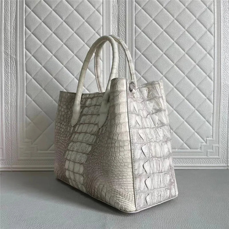 A structured tote bag in textured crocodile-embossed leather, shown in a light gray color. The bag features dual top handles and is positioned against a white quilted paneled wall.