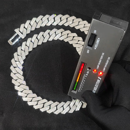 A silver-toned chain necklace with a textured woven or braided link design, displayed on a black fabric background next to a handheld multimeter device with red indicator lights. A black gloved hand is holding the device.