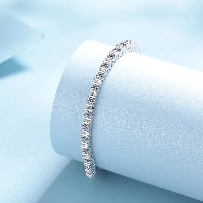 A silver tennis bracelet with baguette-cut clear stones, displayed on a light blue cylindrical jewelry stand against a soft light blue background.