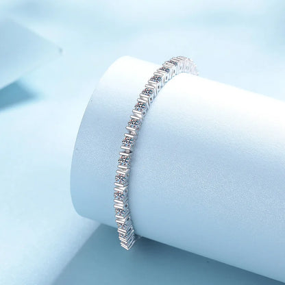 A silver tennis bracelet with baguette-cut clear stones, displayed on a light blue cylindrical jewelry stand against a soft light blue background.