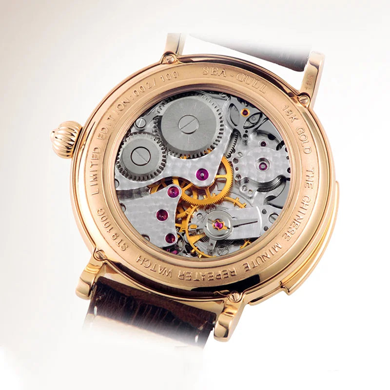 A close-up shot of a gold-tone automatic watch with its back cover removed, showcasing the intricate mechanical movement with silver gears, yellow gold balance wheel, and small pink ruby jewels. The watch has a dark brown leather strap and is set against a clean white background.