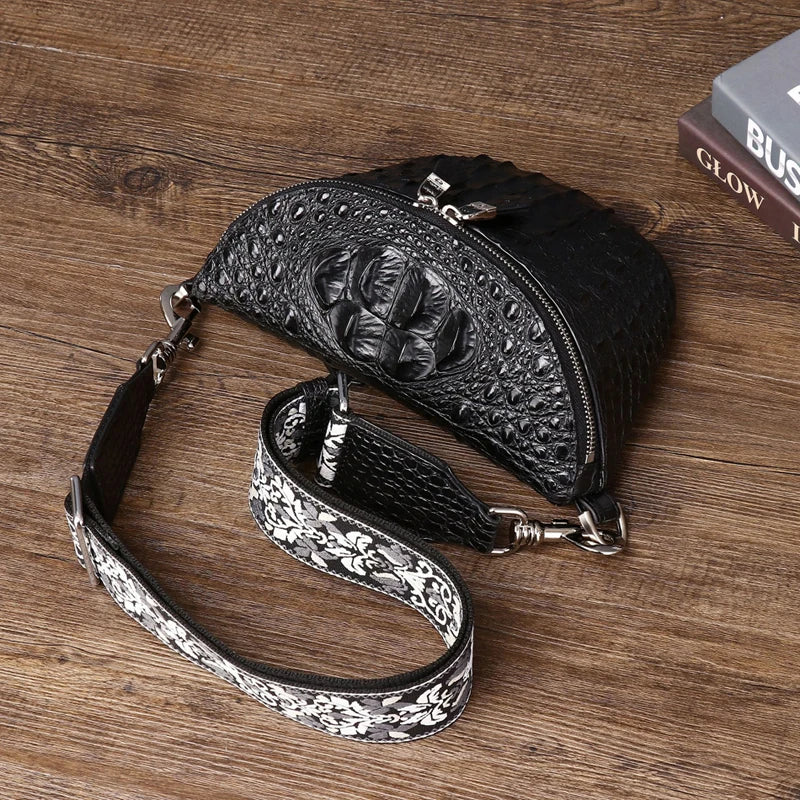 Black crocodile-embossed leather crossbody bag with a patterned strap, displayed on a wooden surface next to a book.