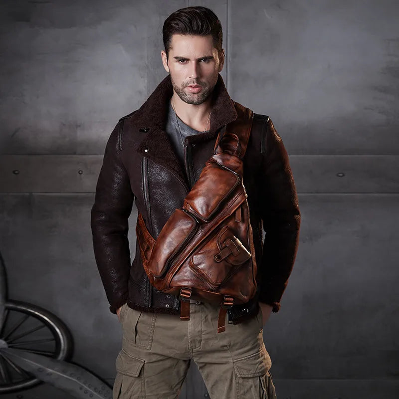 A man wearing a dark brown leather aviator jacket and khaki cargo pants, carrying a large brown leather crossbody sling bag across his chest, set against a dark gray textured wall.