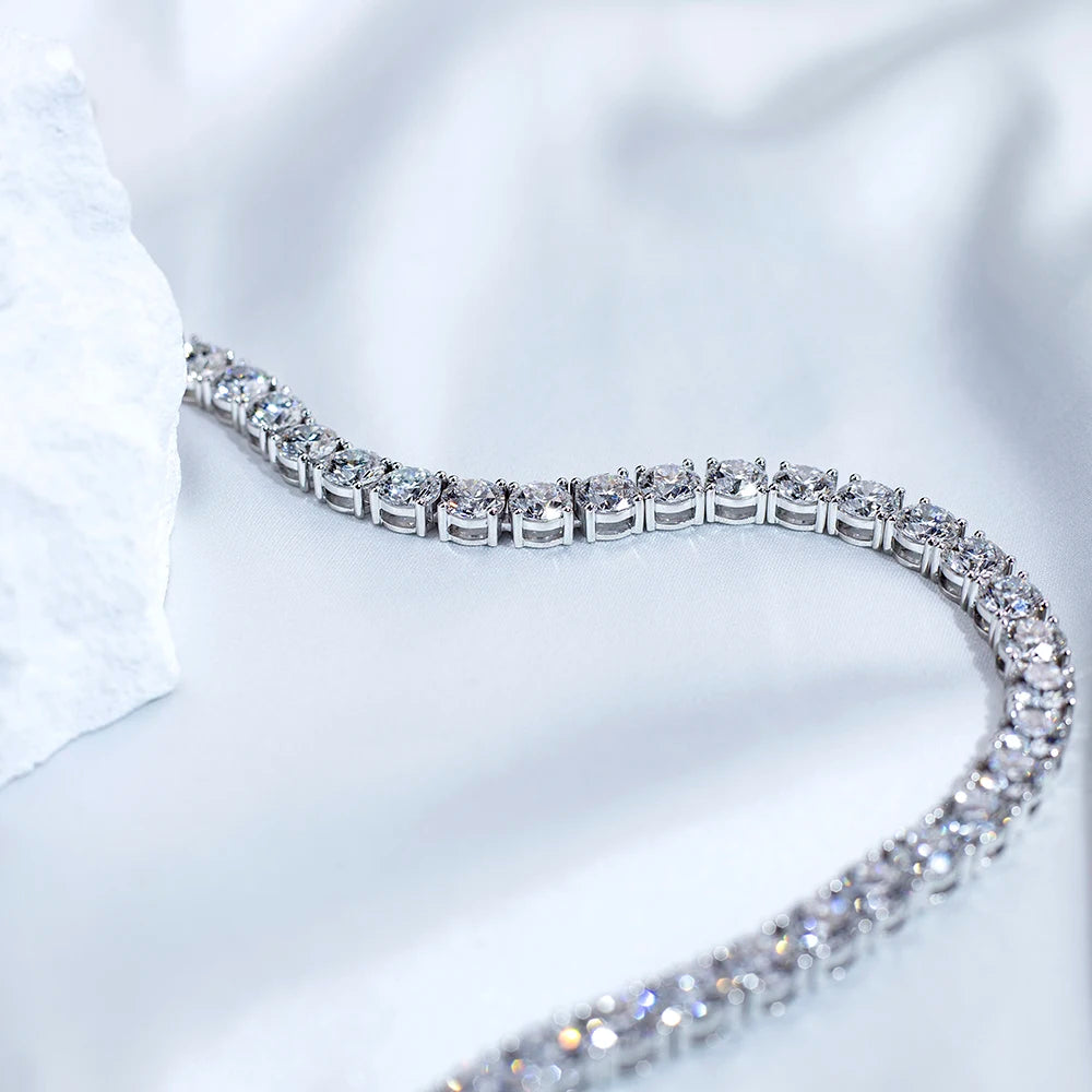 A close-up of a silver tennis bracelet with clear gemstones, arranged in a curved line on a soft white fabric background with a textured white stone prop on the left.