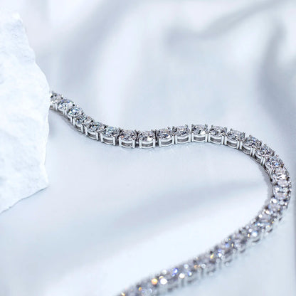 A close-up of a silver tennis bracelet with clear gemstones, arranged in a curved line on a soft white fabric background with a textured white stone prop on the left.