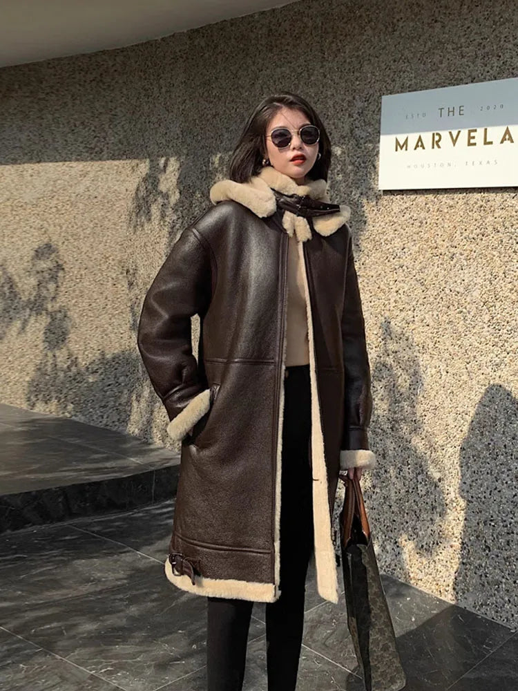 A woman wearing a dark brown shearling-lined leather coat with a beige fur-trimmed collar, standing outdoors against a textured stone wall.