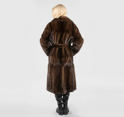 A person shown from the back wearing a long brown fur coat with a matching belt, paired with black boots, against a light gray studio background.