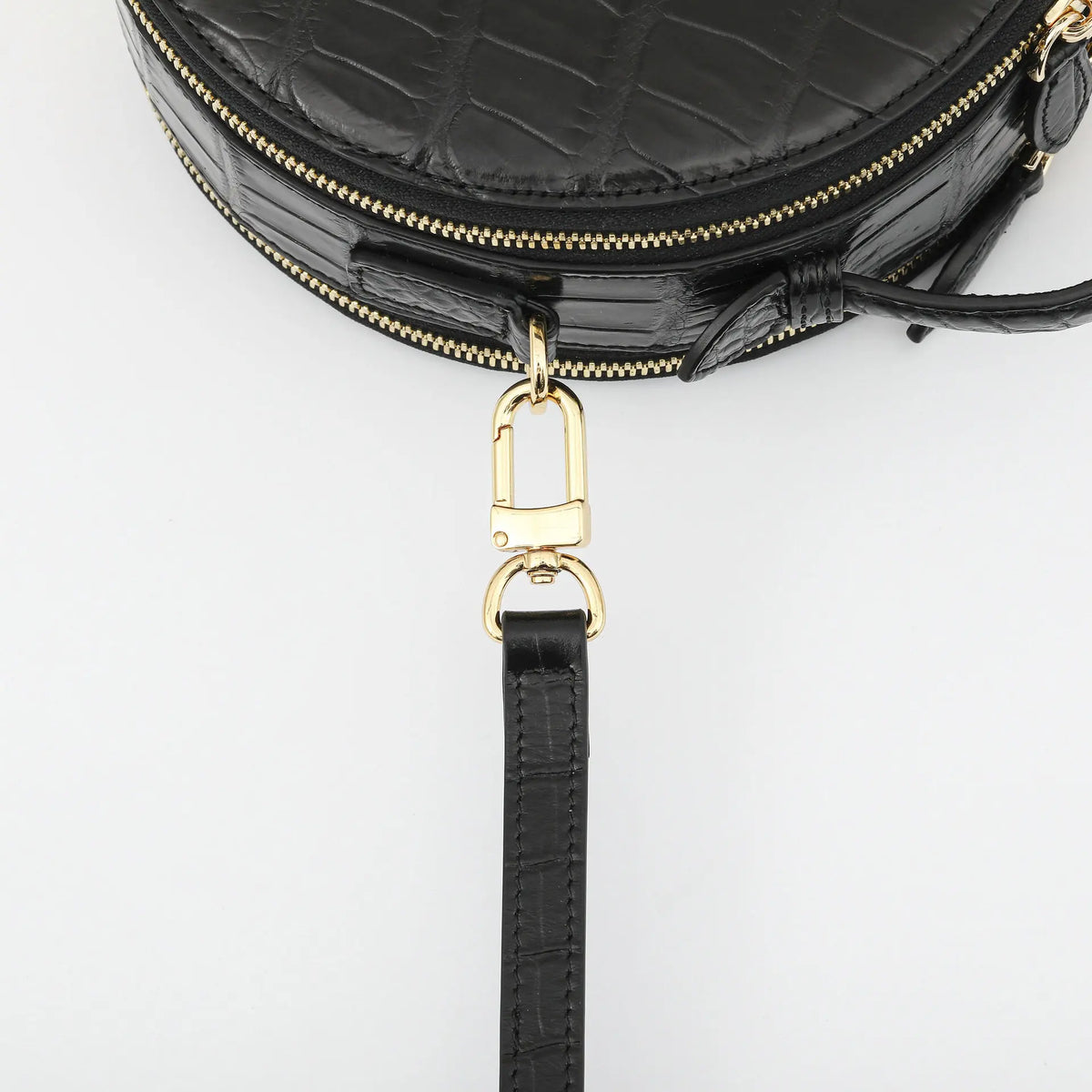 Close-up of a black crocodile-embossed leather handbag with gold-tone hardware and a detachable black leather wristlet strap, shown on a light gray background.