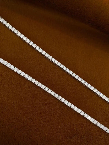 A close-up of a silver tennis chain necklace with clear rectangular gemstones, laid diagonally on a textured brown surface.