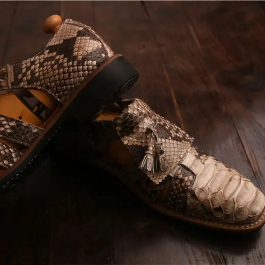 A pair of men's snakeskin leather shoes with cutout straps and tassel details, displayed on a dark wooden floor.
