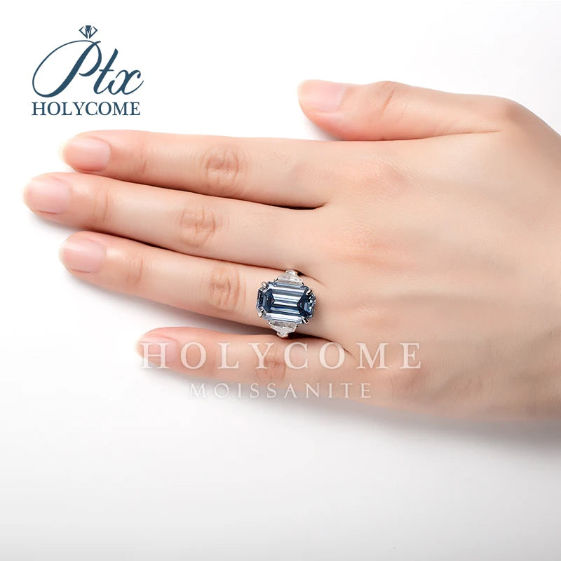 A hand wearing a silver ring with a large emerald-cut blue moissanite center stone, shown against a clean white background with the brand logo 'Plc HOLYCOME' and 'HOLYCOME MOISSANITE' text overlays.