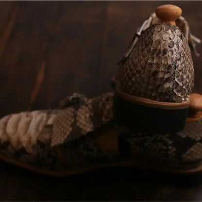 A close-up of a pair of snakeskin-patterned sandals resting on a dark wooden surface, with one sandal standing upright to show the sole and heel detail.