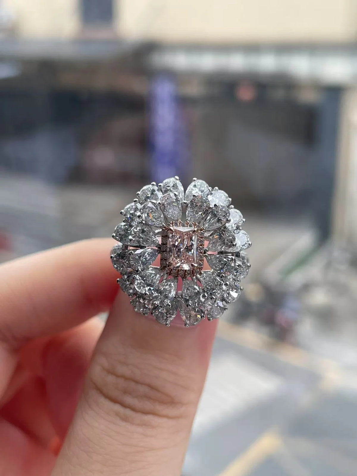 A close-up of a hand holding a large diamond halo ring with a central pink gemstone, set against a blurred outdoor street background.