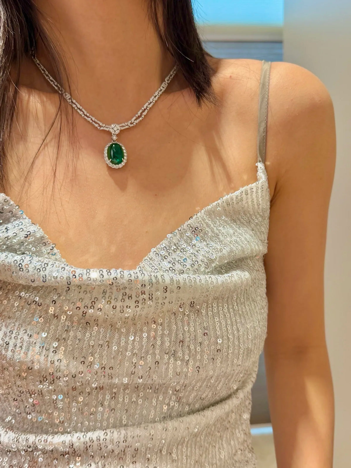 A close-up of a model wearing a silver chain necklace with a green oval pendant, paired with a silver sequined camisole top.