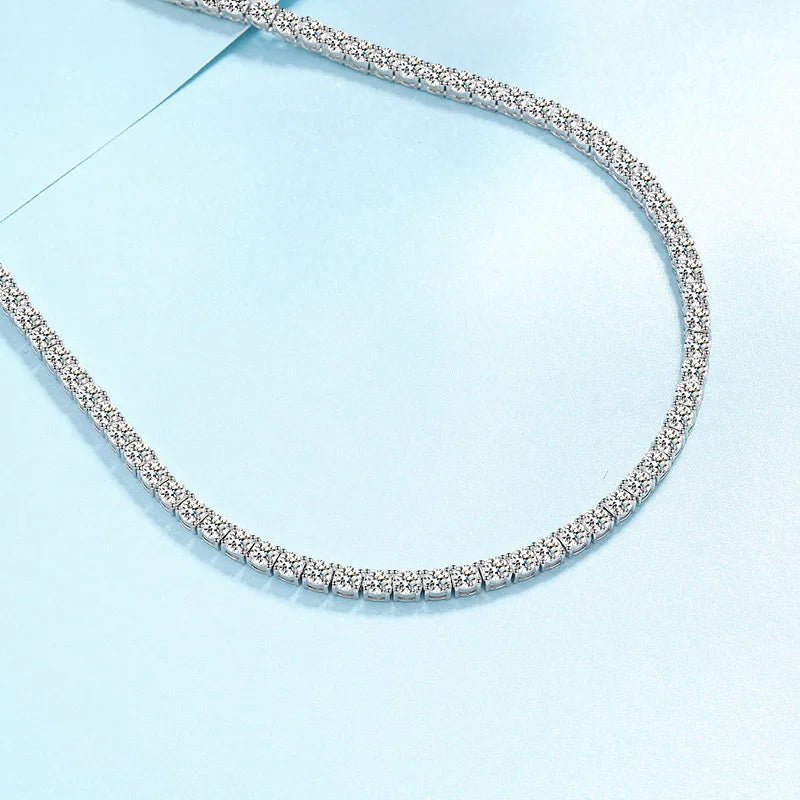 A silver tennis necklace paved with small clear stones, arranged in a curved loop on a light blue background.