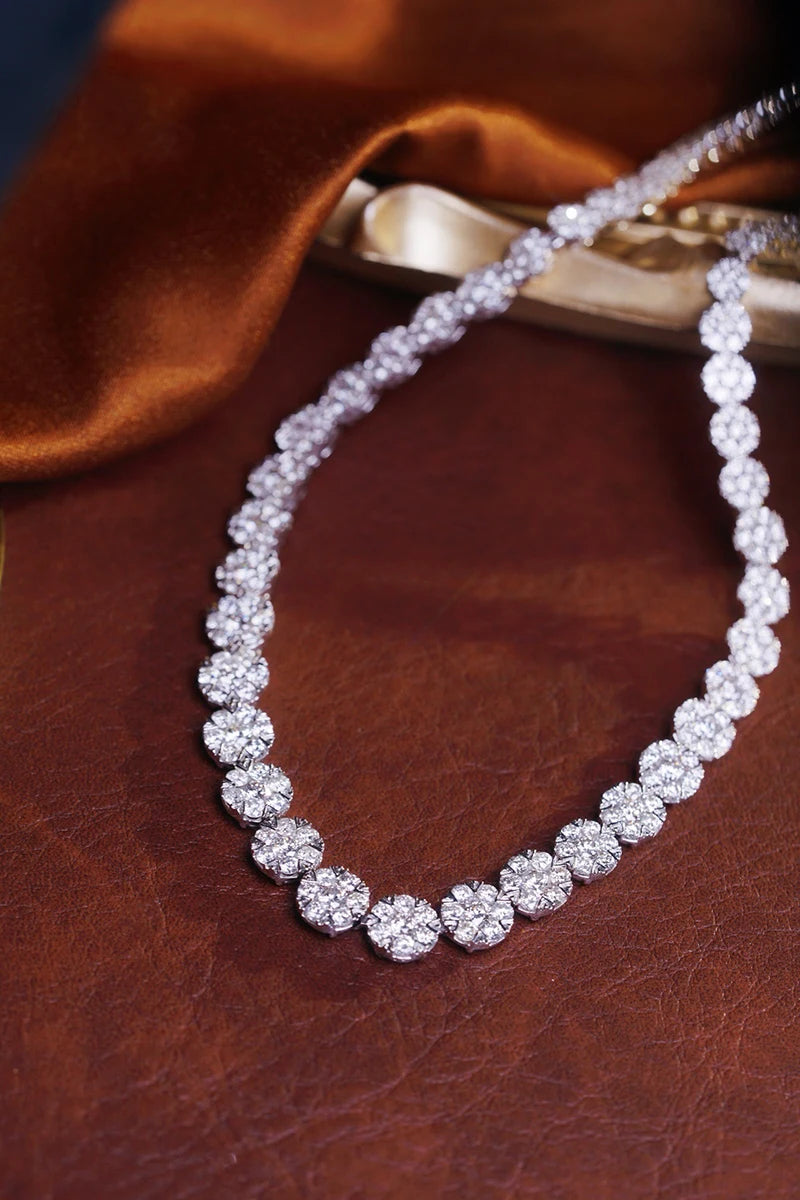 A diamond tennis necklace with round floral clusters of clear stones set in silver-toned metal, displayed on a textured brown leather surface with a draped brown satin fabric in the background.