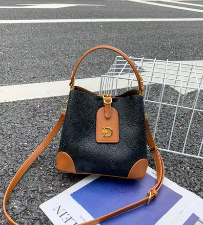A small black embossed bucket bag with tan leather trim, a top handle, and a detachable shoulder strap, placed on asphalt over a magazine page and wire crate.