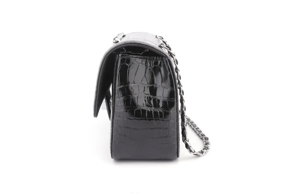 A black crocodile-embossed leather shoulder bag with a glossy finish and a silver chain strap, shown from a side profile view on a white background.