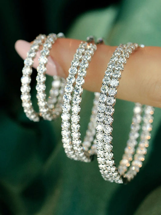 Close-up of a hand wearing multiple silver-toned tennis bracelets adorned with sparkling clear stones, set against a dark green blurred background.