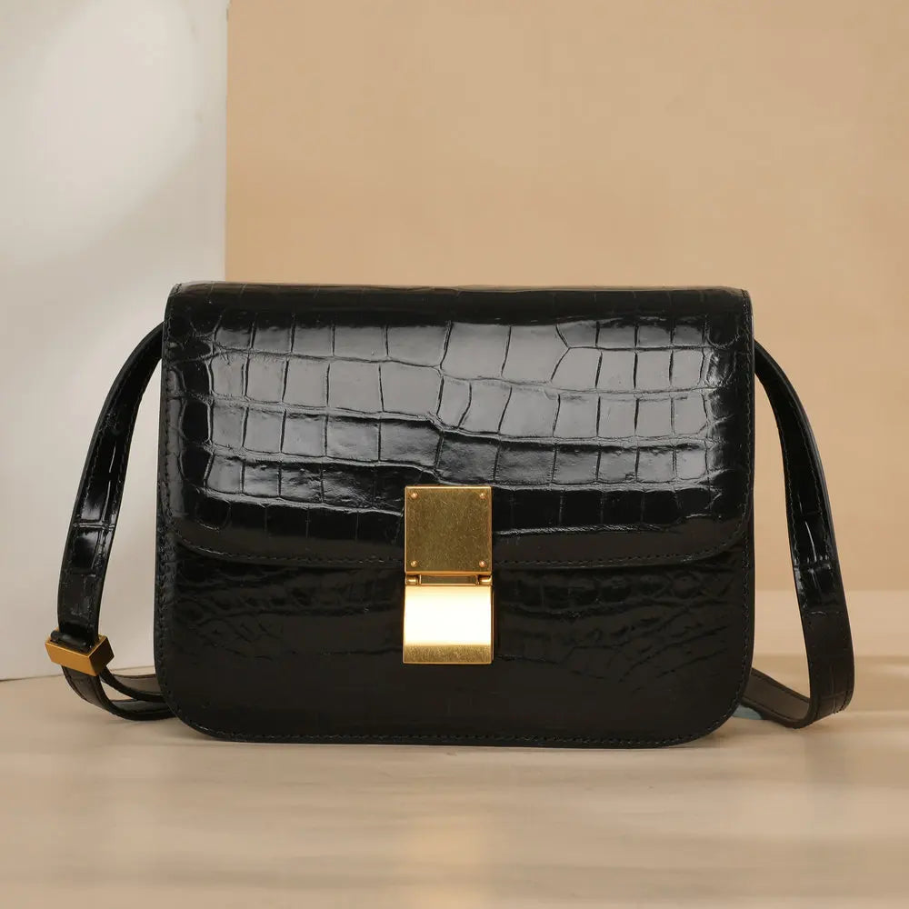 A black crocodile-embossed leather shoulder bag with a gold-tone metal clasp and a thin black strap, set against a neutral beige background.