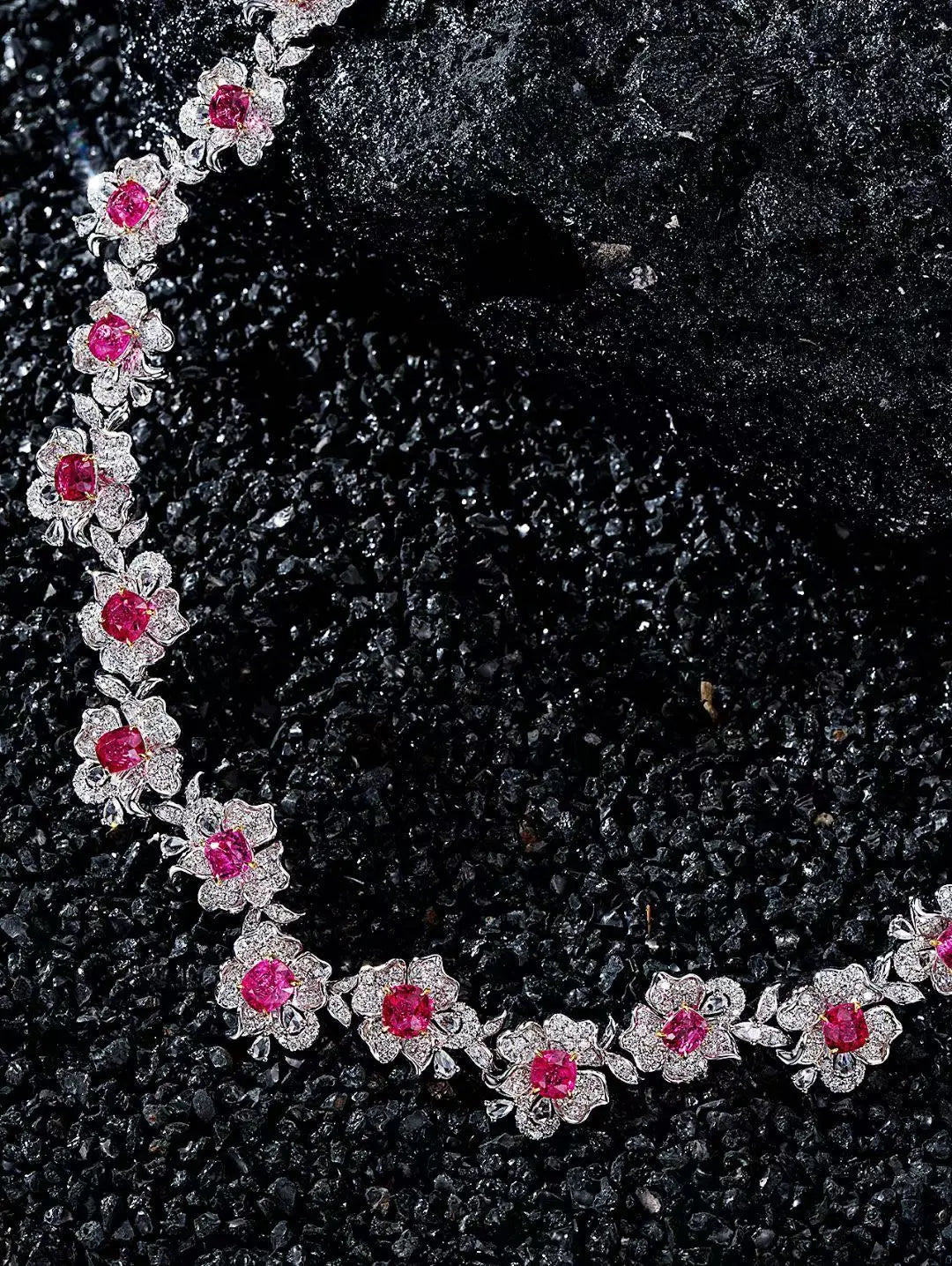 A close-up of a floral necklace with bright pink gemstones and silver-toned settings, arranged on a dark textured surface.