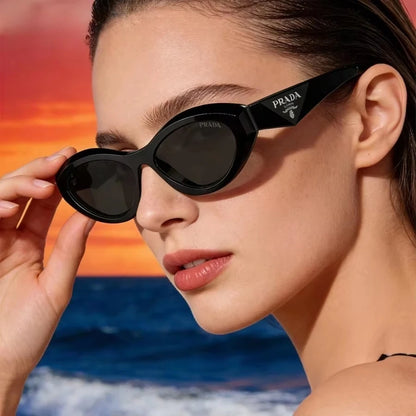A close-up lifestyle shot of a model wearing black Prada cat-eye sunglasses against a vibrant beach sunset background.