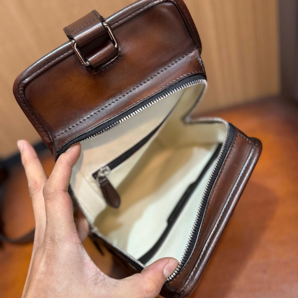 A close-up of a brown leather pouch or small bag being held open by a hand, showing a cream-colored interior lining, zipper closure, and metal buckle detail.