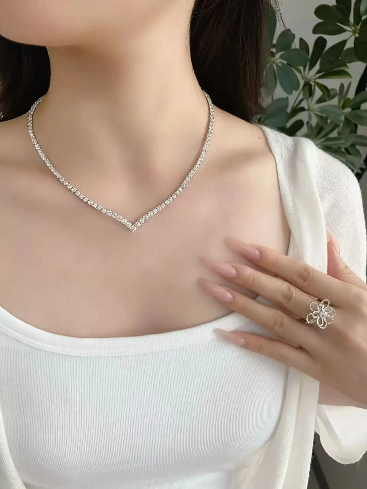 A close-up of a woman wearing a delicate silver V-shaped necklace with clear stones, paired with a white top and a matching silver flower ring on her finger.
