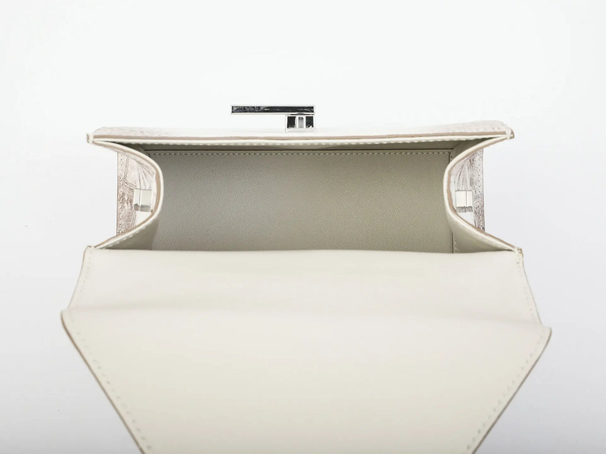 An open cream-colored leather clutch bag with a structured geometric shape, featuring a light beige interior lining and a silver-tone metal clasp at the top, set against a plain white background.