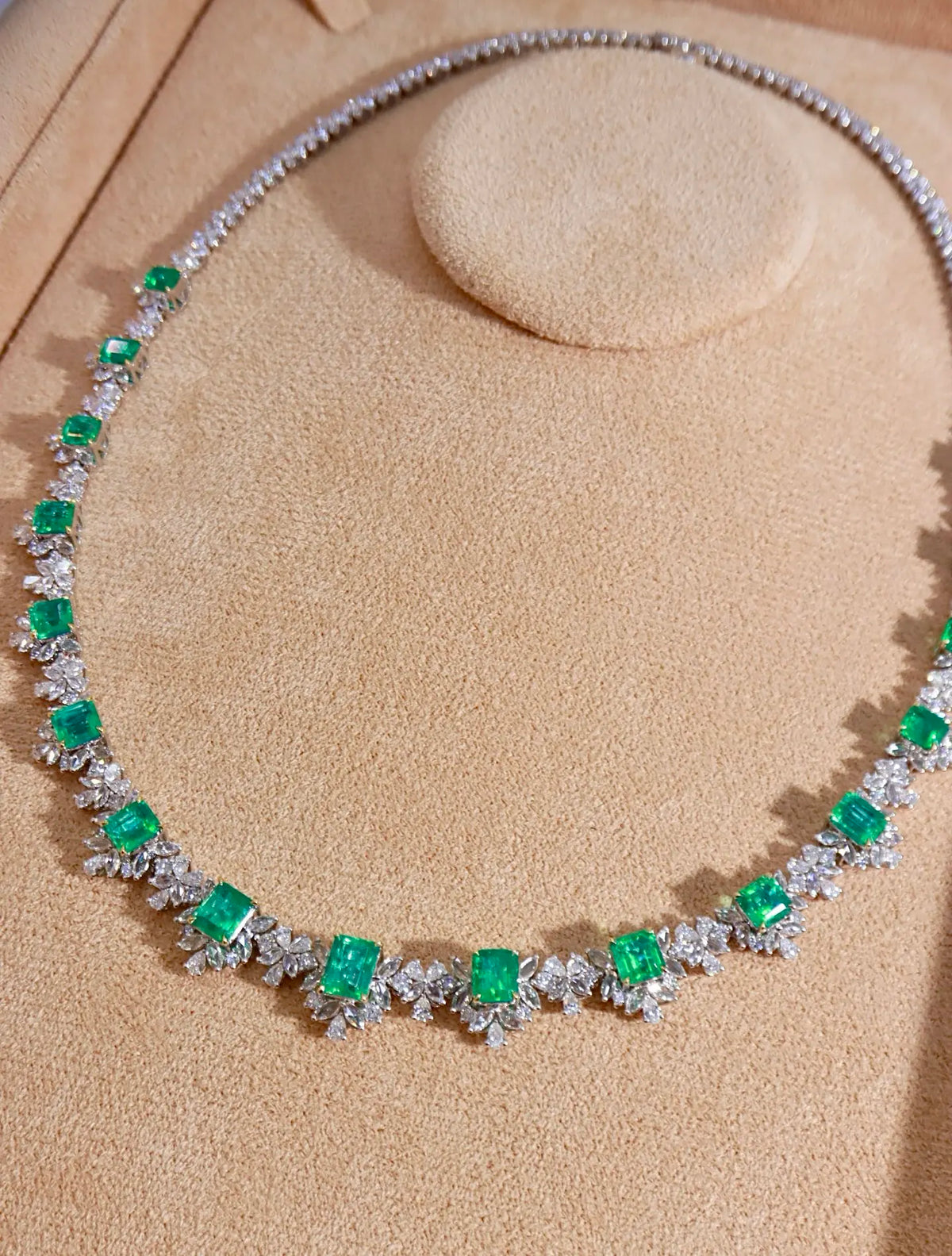 A silver-toned necklace featuring square-cut green emeralds and pavé-set clear stones, displayed on a beige jewelry bust.
