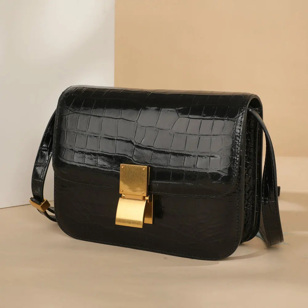 A black crocodile-embossed leather shoulder bag with a prominent gold-tone clasp and a matching black strap, set against a minimalist beige and white background.