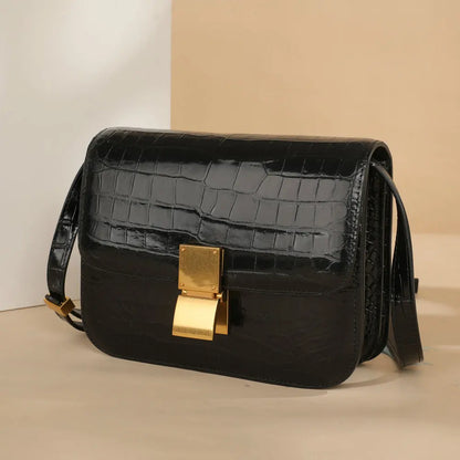 A black crocodile-embossed leather shoulder bag with a prominent gold-tone clasp and a matching black strap, set against a minimalist beige and white background.