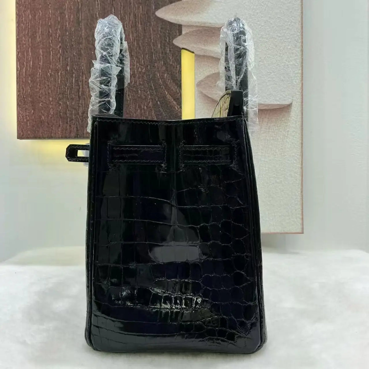 A glossy black crocodile-embossed leather handbag with protective plastic wrapping on its handles, displayed against a neutral indoor background.