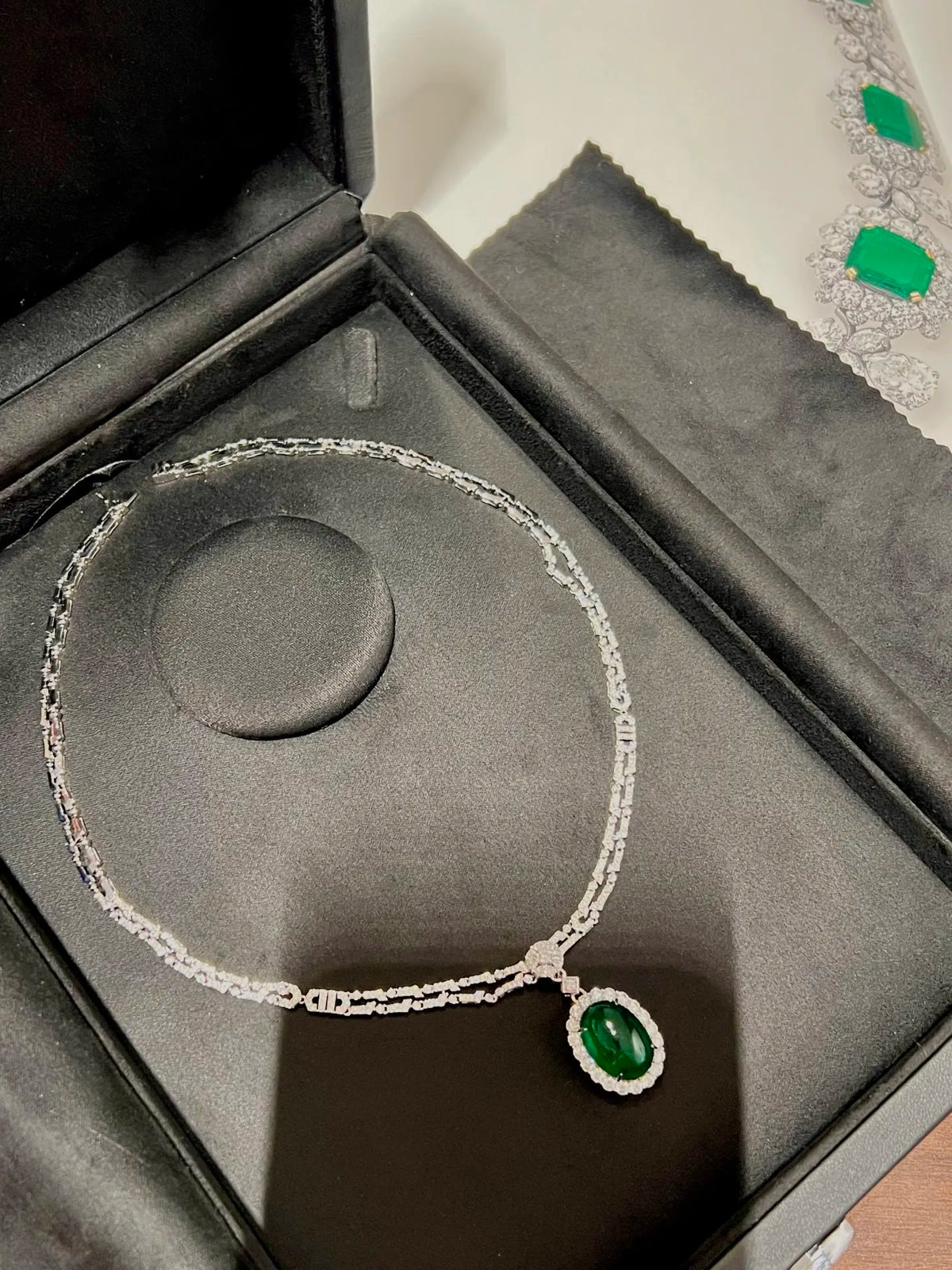 A silver-toned chain necklace with a green oval pendant, displayed inside an open black jewelry box with a gray velvet insert.