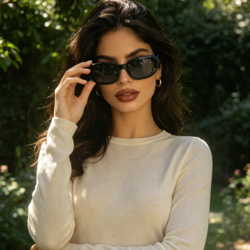 A woman outdoors wearing black cat-eye sunglasses and a cream-colored long-sleeve knit top, with a blurred green garden background.