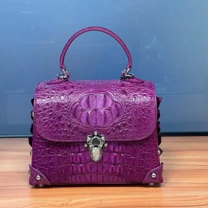 A structured purple handbag with a crocodile-embossed texture, featuring a top handle, silver-tone hardware, and decorative studs, set against a blue background.