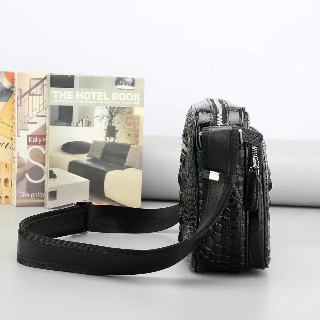 A black crocodile-embossed leather crossbody bag with a matching adjustable shoulder strap, displayed next to a book on a light gray surface.