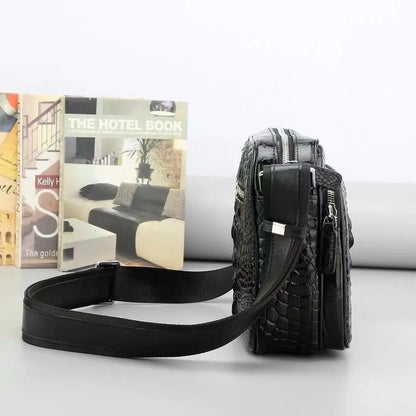A black crocodile-embossed leather crossbody bag with a matching adjustable shoulder strap, displayed next to a book on a light gray surface.