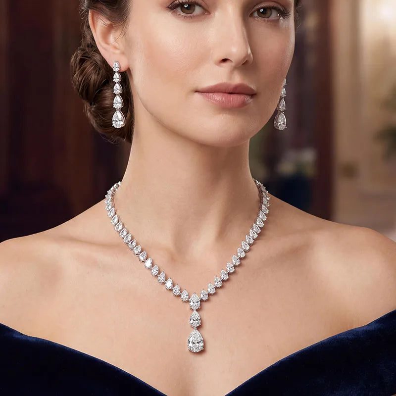 A close-up of a woman wearing a sparkling diamond necklace with a teardrop-shaped pendant and matching drop earrings, set against a softly blurred warm-toned indoor background.