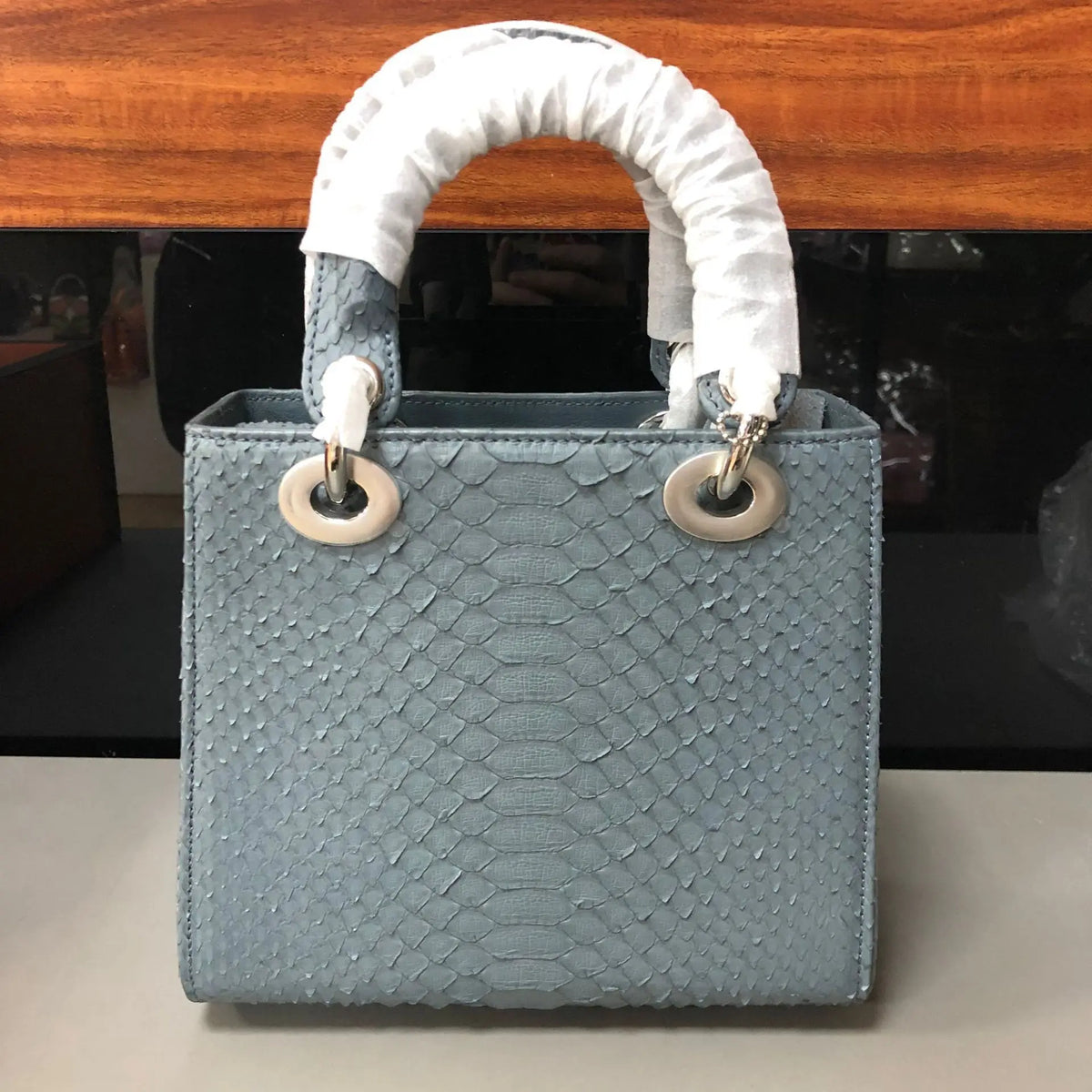 A structured light blue handbag with a textured python-style embossed pattern, featuring white protective wrapping on the handles and silver-tone hardware.